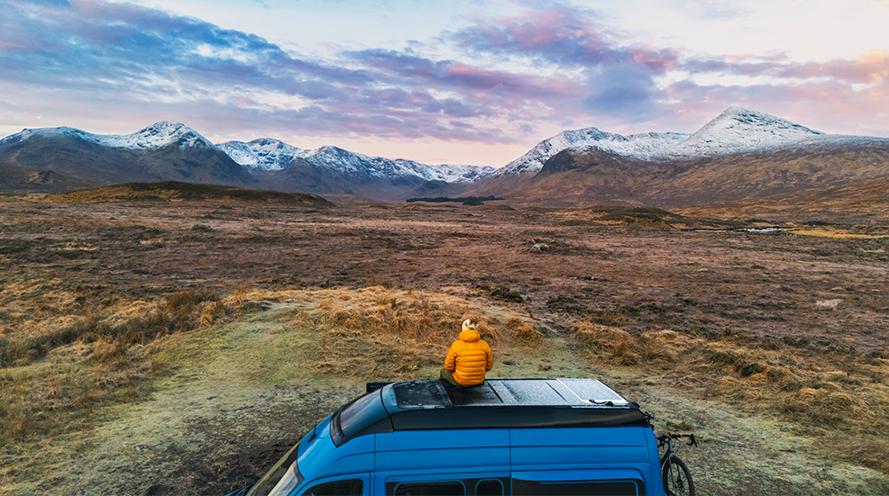 Person sat on top of van looking out at view of snowy mountain tops